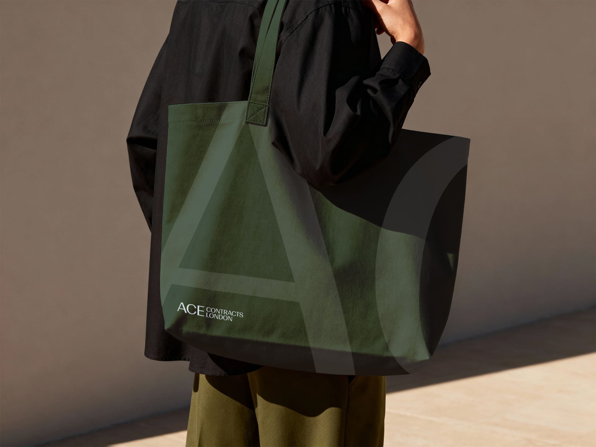 Person holding a dark green tote bag with “ACE CONTRACTS LONDON” and large stylized “AC” letters printed on it, standing against a neutral background.