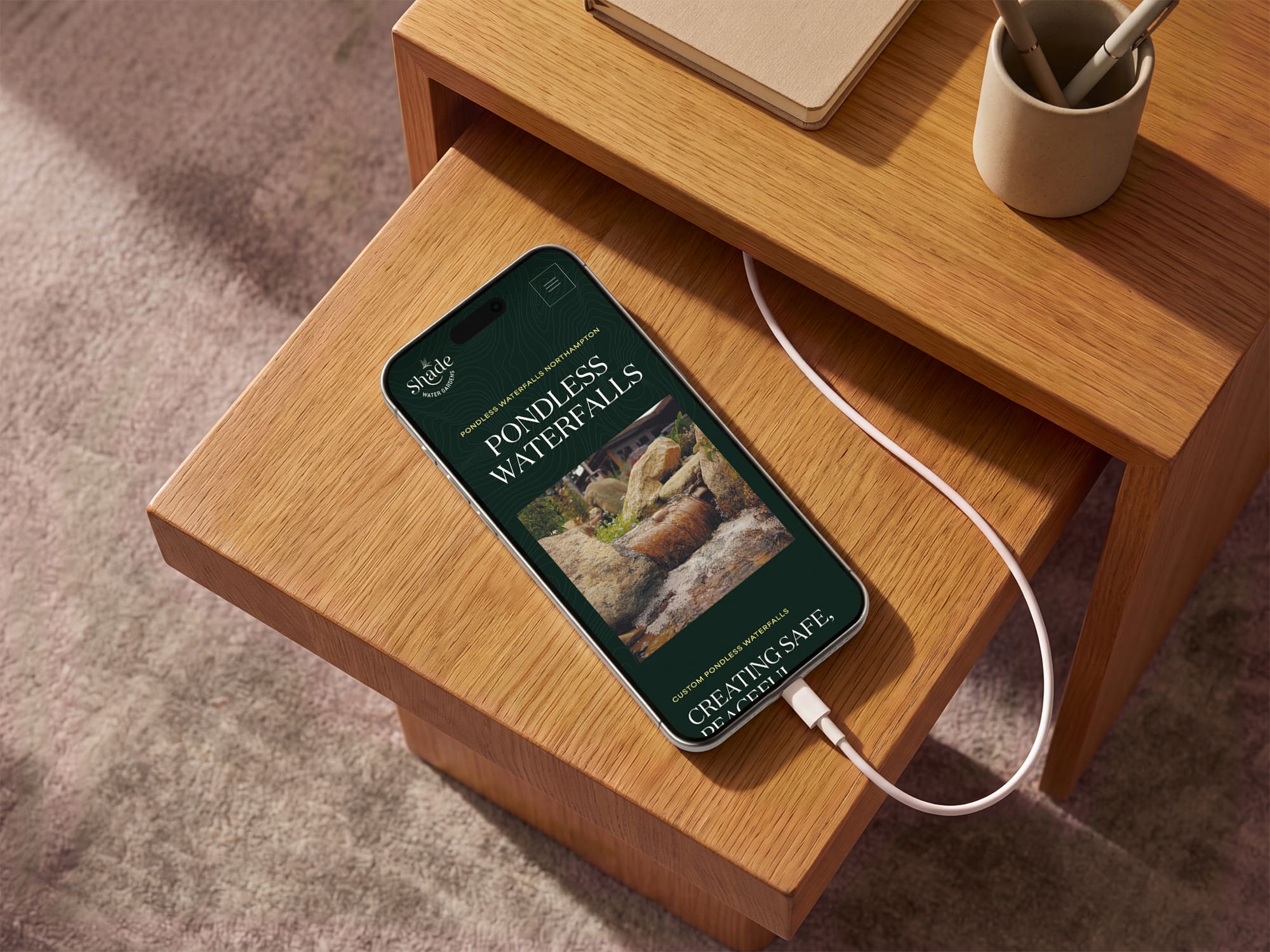 A smartphone charging on a wooden table displays a webpage about pondless waterfalls, featuring a photo of a small waterfall and the text “Creating Safe, Peaceful Spaces.”.