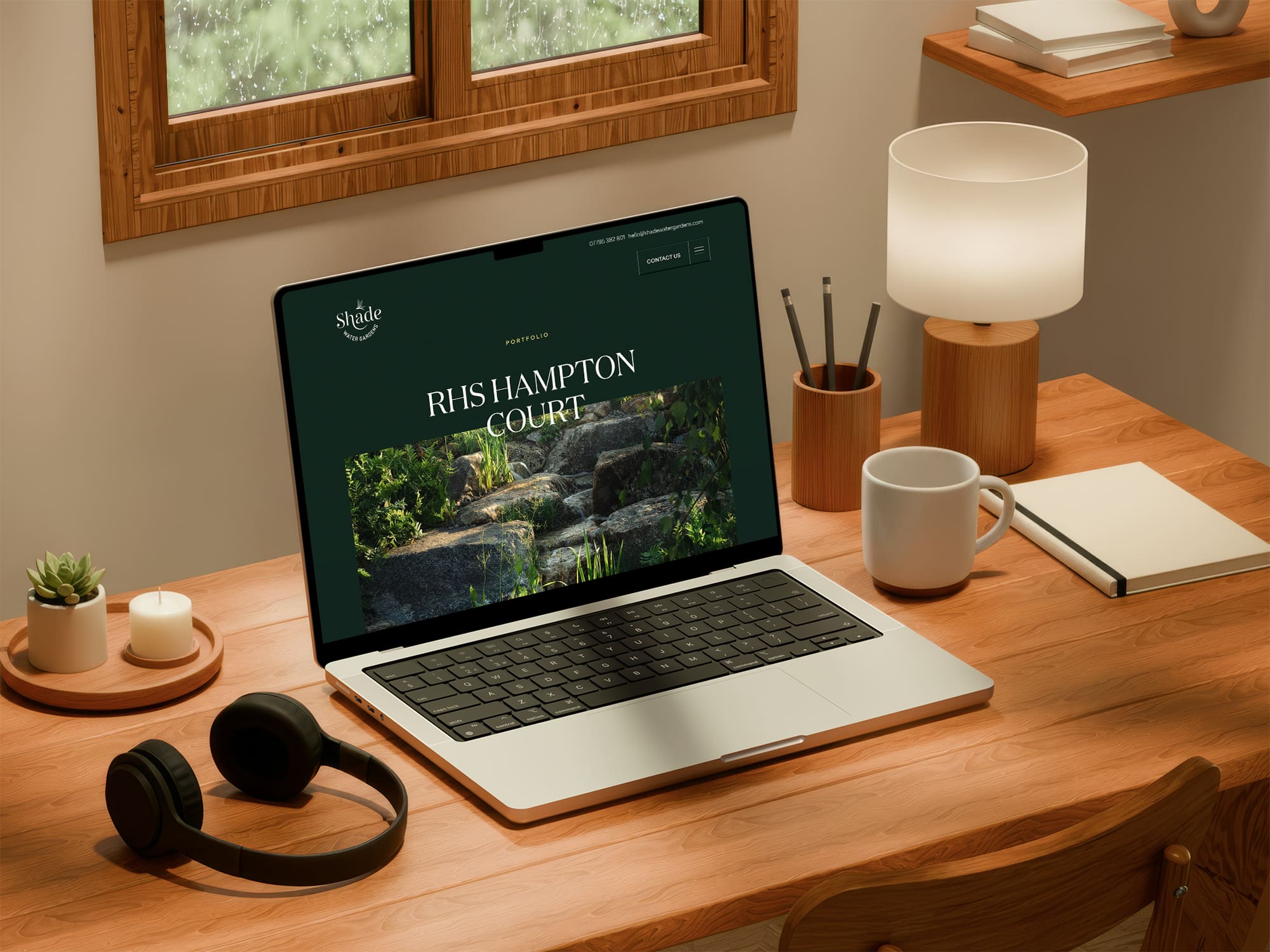 A laptop on a wooden desk displays a website with the heading “RHS Hampton Court.” The desk also holds headphones, a lamp, mugs, pens, and notebooks near a window.