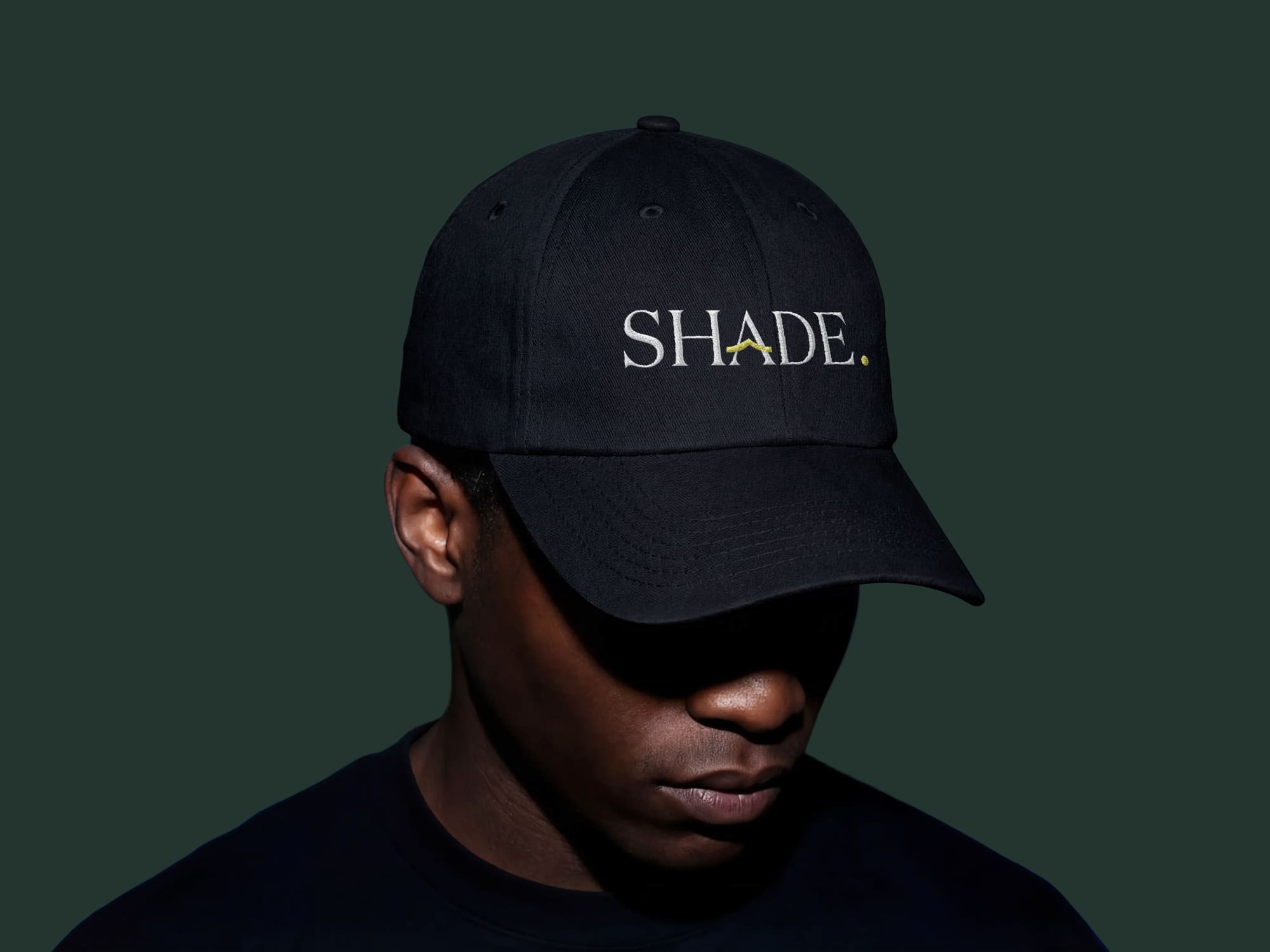 A person wearing a black cap with the word “SHADE.” embroidered on it in white and yellow lettering, set against a dark green background.